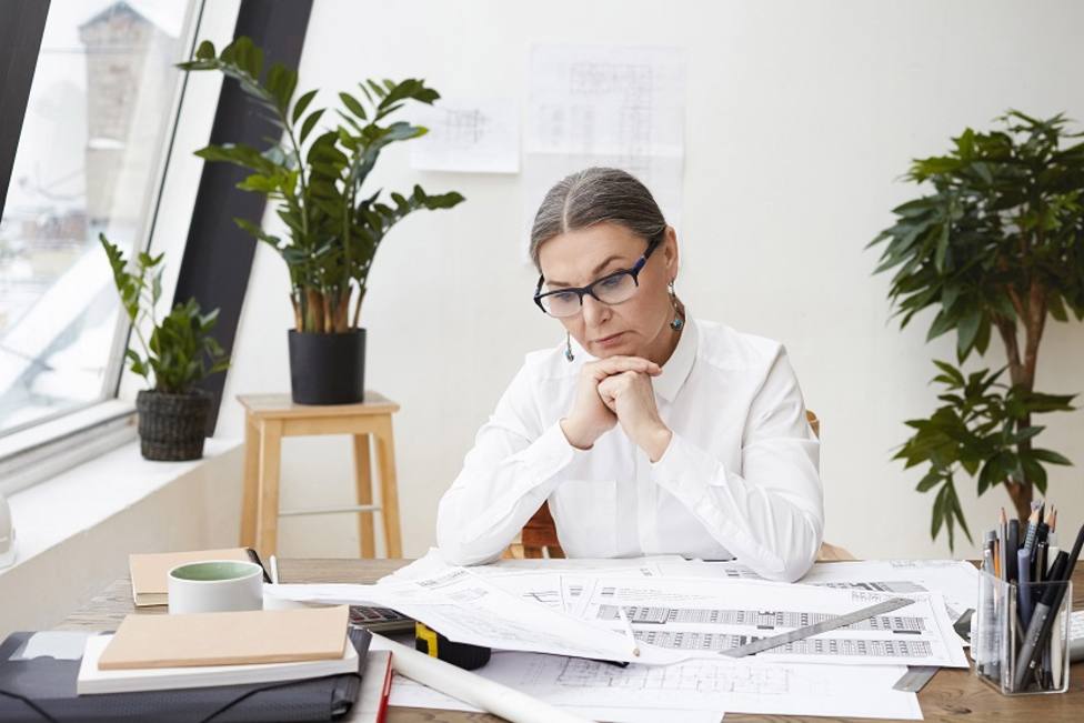 Horizontal shot of pensive middle aged woman engineer wearing black glasses and white shirt holding clasped hands under her chin, studying drawings and specifications on desk in front of her